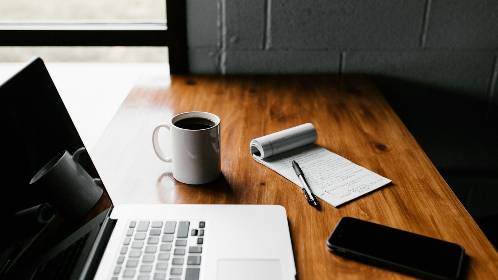 A laptop, coffee, notepad, and phone on a wooden desk. Photograph by Andrew Neel, Unsplash.
