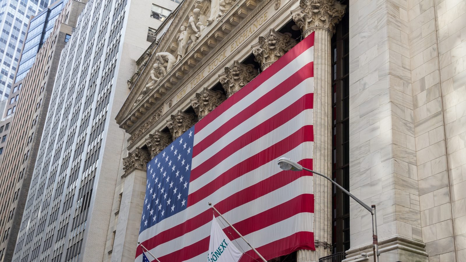 The New York Stock Exchange on Wall Street, with a large American flag on the facade. Photograph by Dietmar Rabich, CC BY-SA.