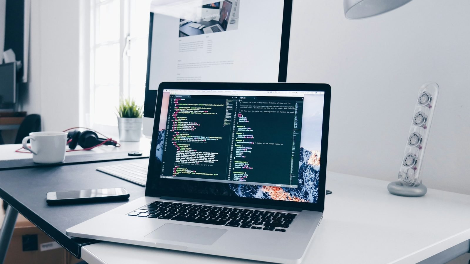 A silver laptop, phone, mug, and headphones on a white desk. Photograph by Christopher Gower, Unsplash.