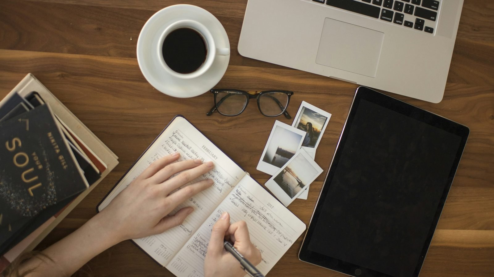 Hands writing in a notebook on a wooden desk beside a laptop, coffee, and glasses. Photograph by Tim Gouw, Unsplash.